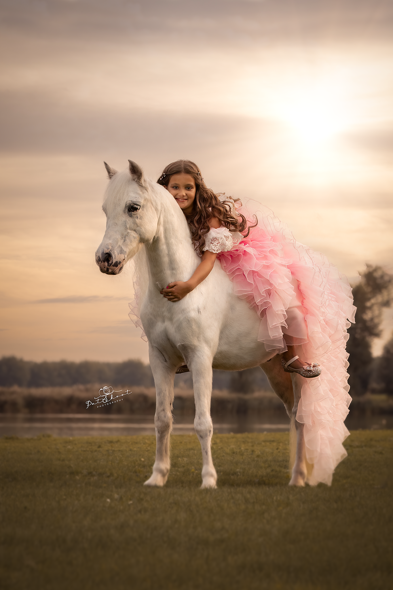 Meisje in een roze jurk rijdt op een witte pony in een grasveld en lacht. Zonsondergang op de achtergrond.