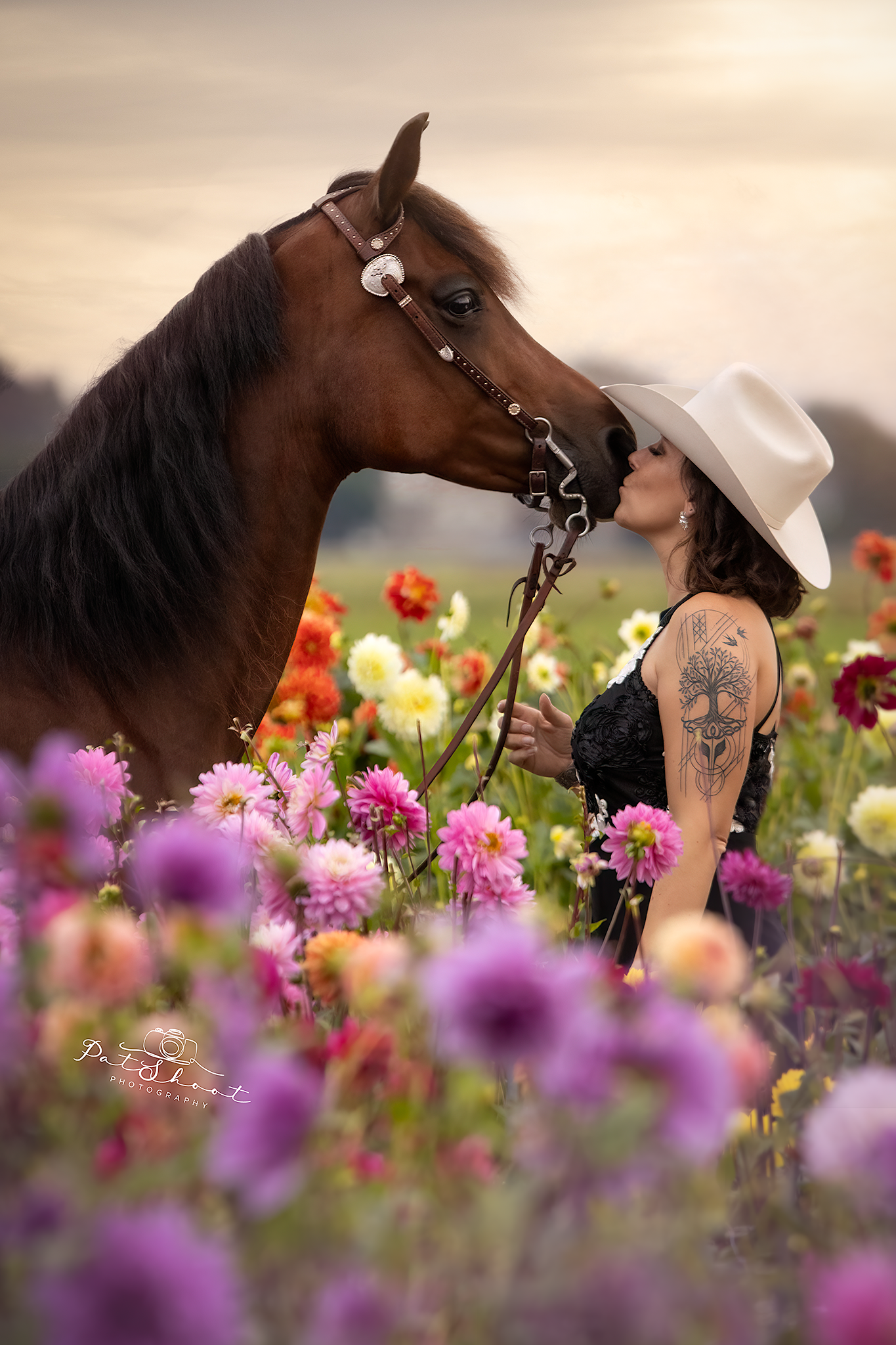 Een vrouw met een cowboyhoed kust de neus van een bruin paard in een veld vol kleurrijke bloemen.