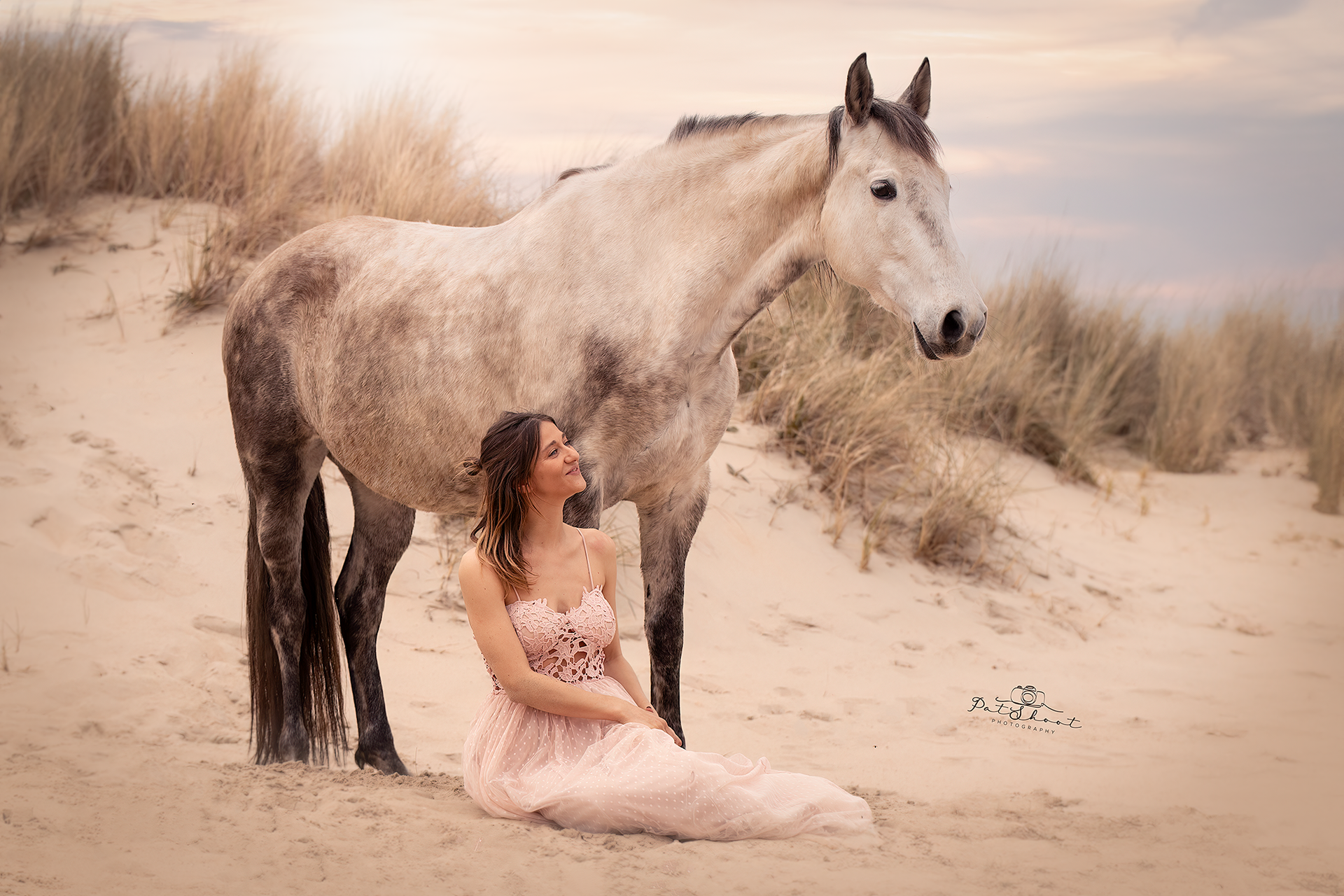 Een vrouw in een roze jurk zit op het zand, naast een wit paard, met duinen op de achtergrond, in zacht licht.