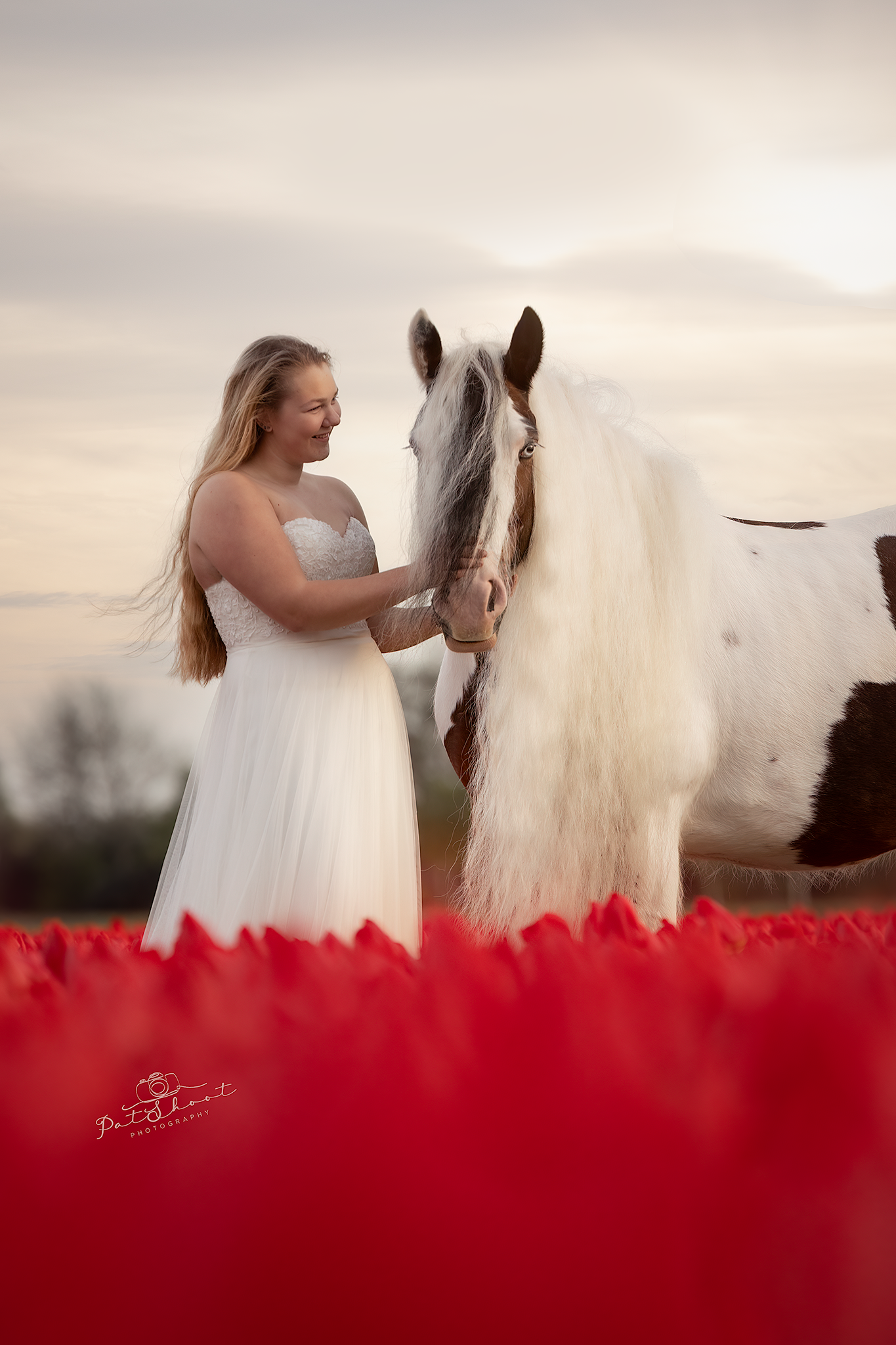 Bruid in witte jurk, die een wit-bruin paard aait in een veld met rode bloemen.