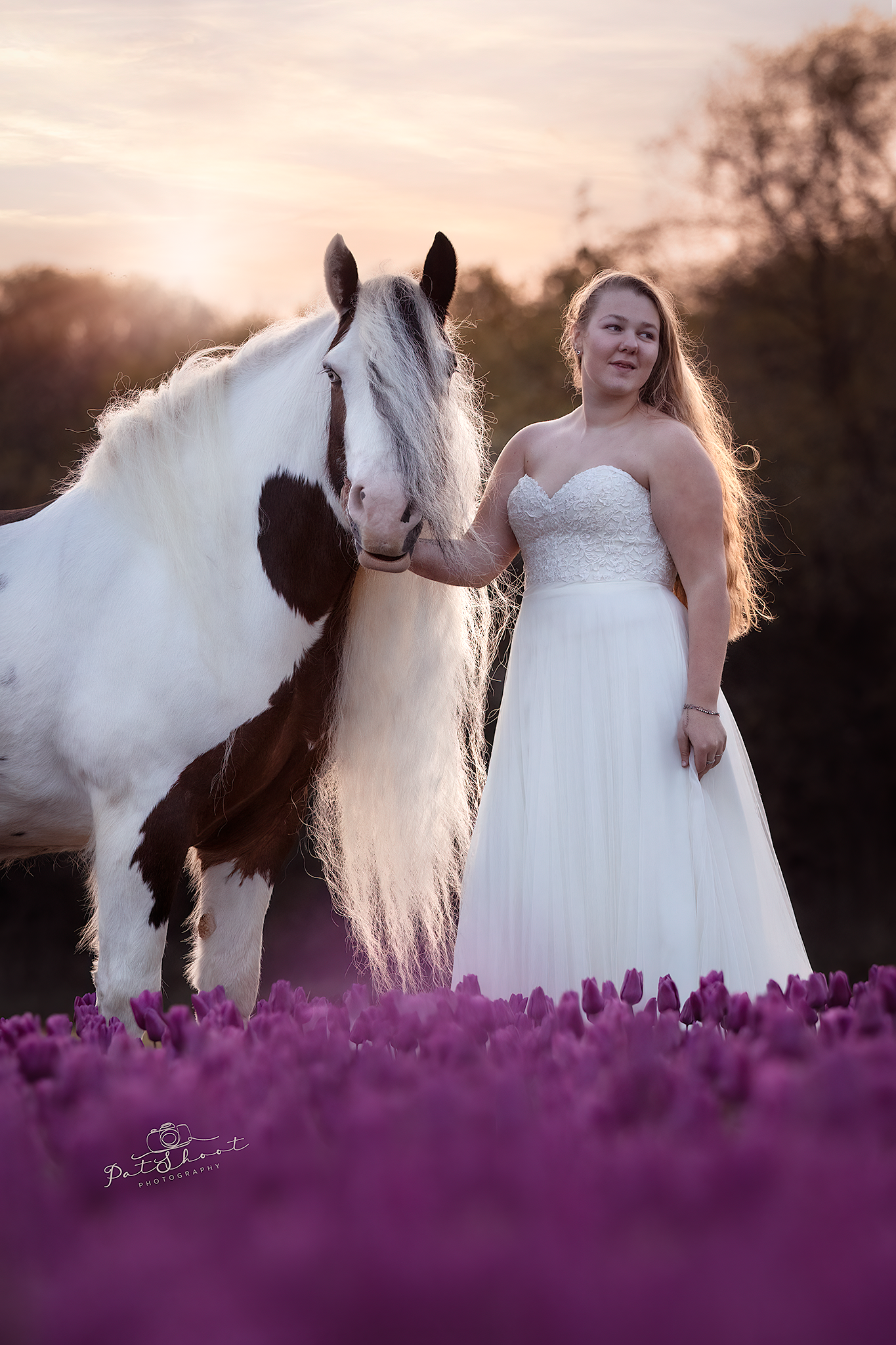 Vrouw in trouwjurk met beschilderd paard in paarse bloemen bij zonsondergang.