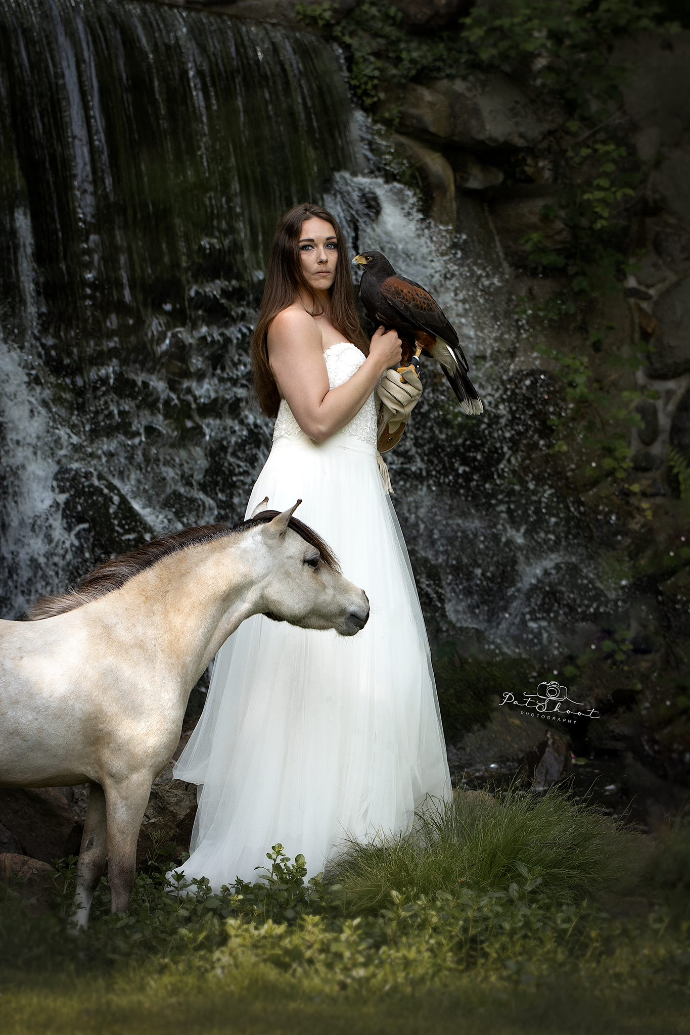 Vrouw in witte jurk met valk staat bij een waterval, pony links van haar.