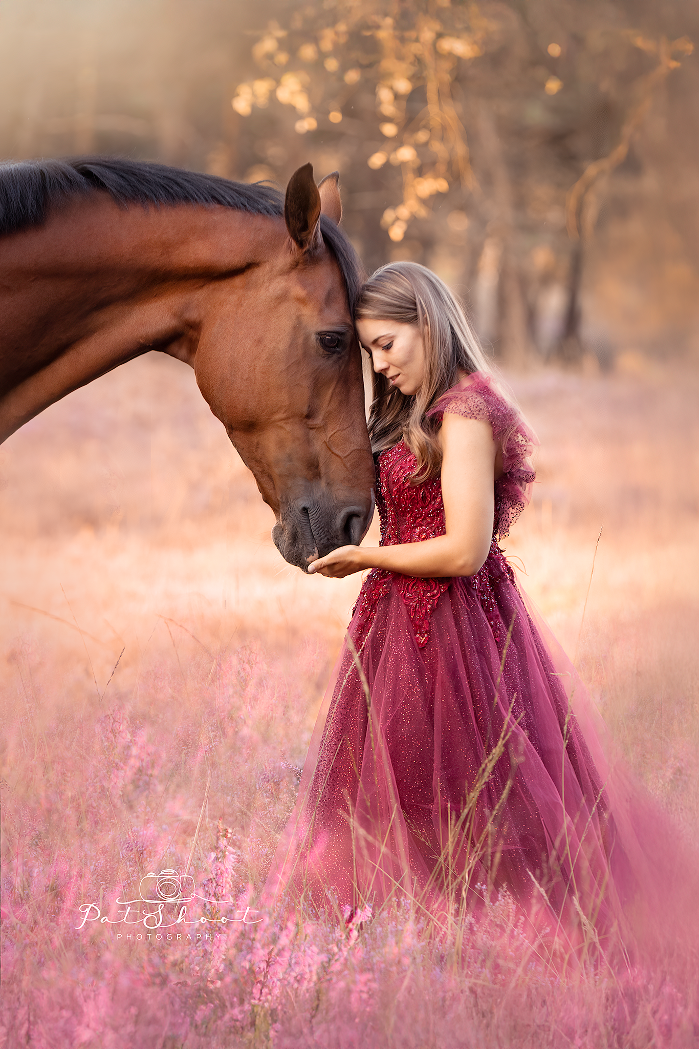 Vrouw in rode jurk met bruin paard in een veld met roze bloemen; hun voorhoofden raken elkaar aan.