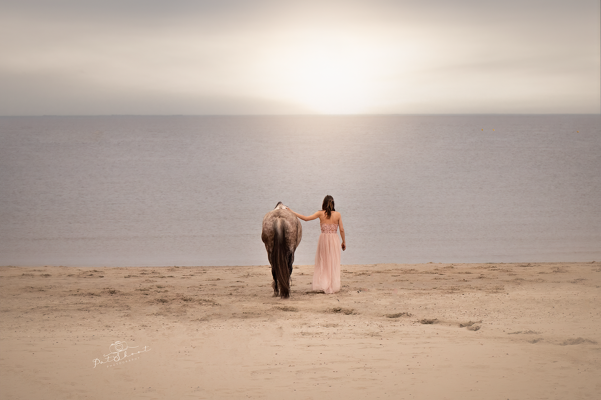 Een vrouw in een roze jurk loopt met een paard over een zandstrand bij zonsondergang.