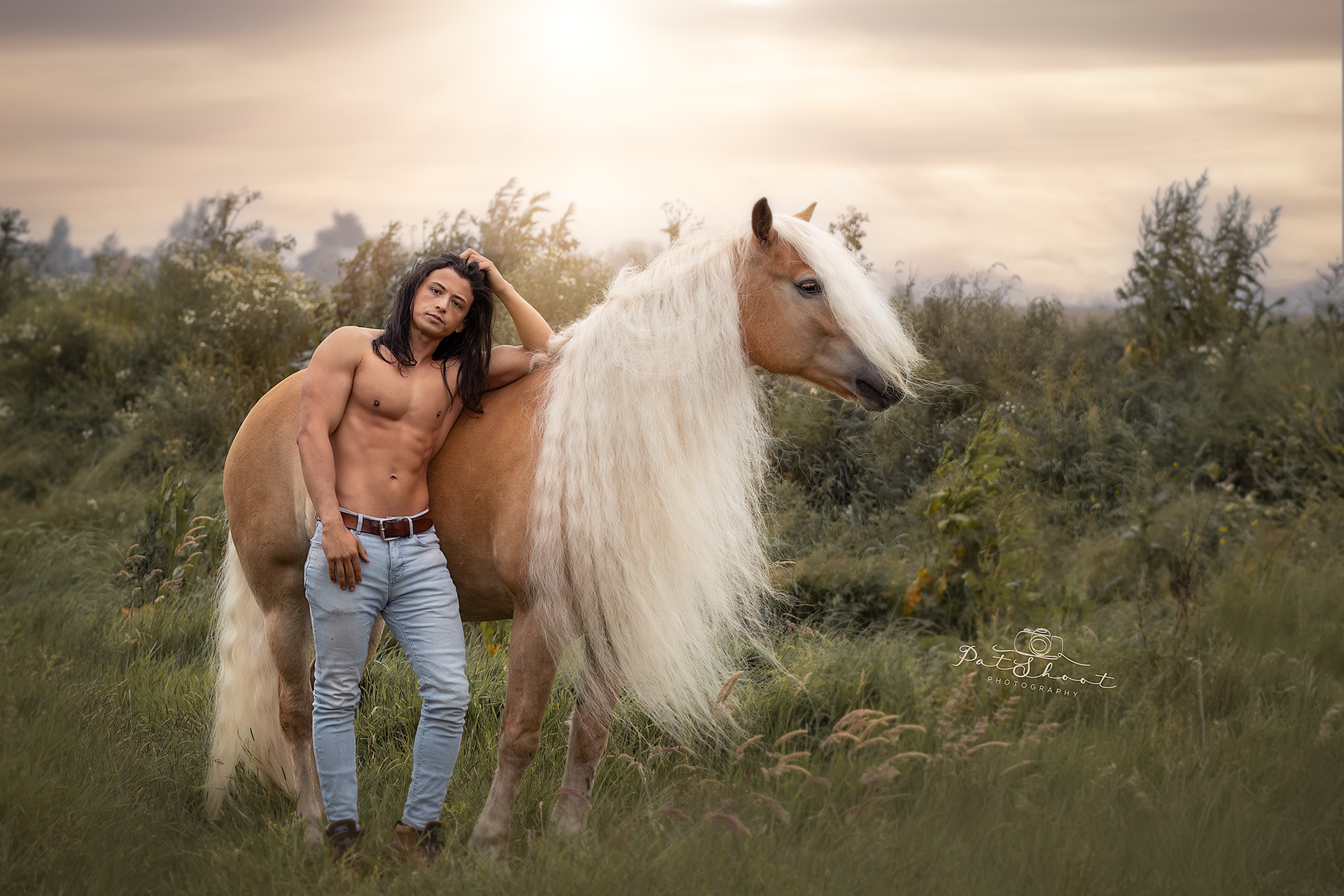 Een man leunt tegen een bruin paard met een lange witte manen in een veld; de zon gaat onder.