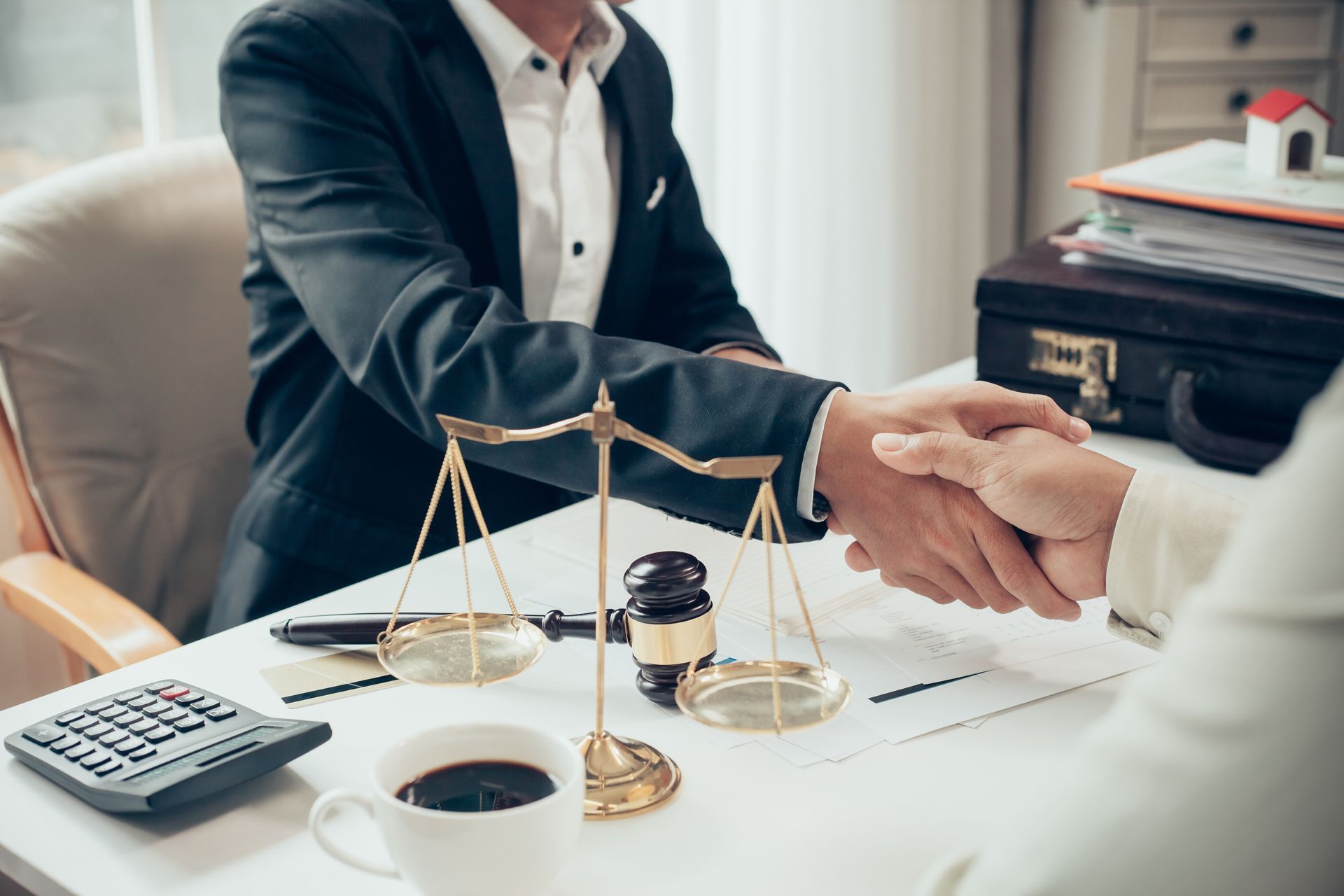 Deux personnes se serrent la main devant un bureau où se trouvent une balance de la justice, du café et une calculatrice.