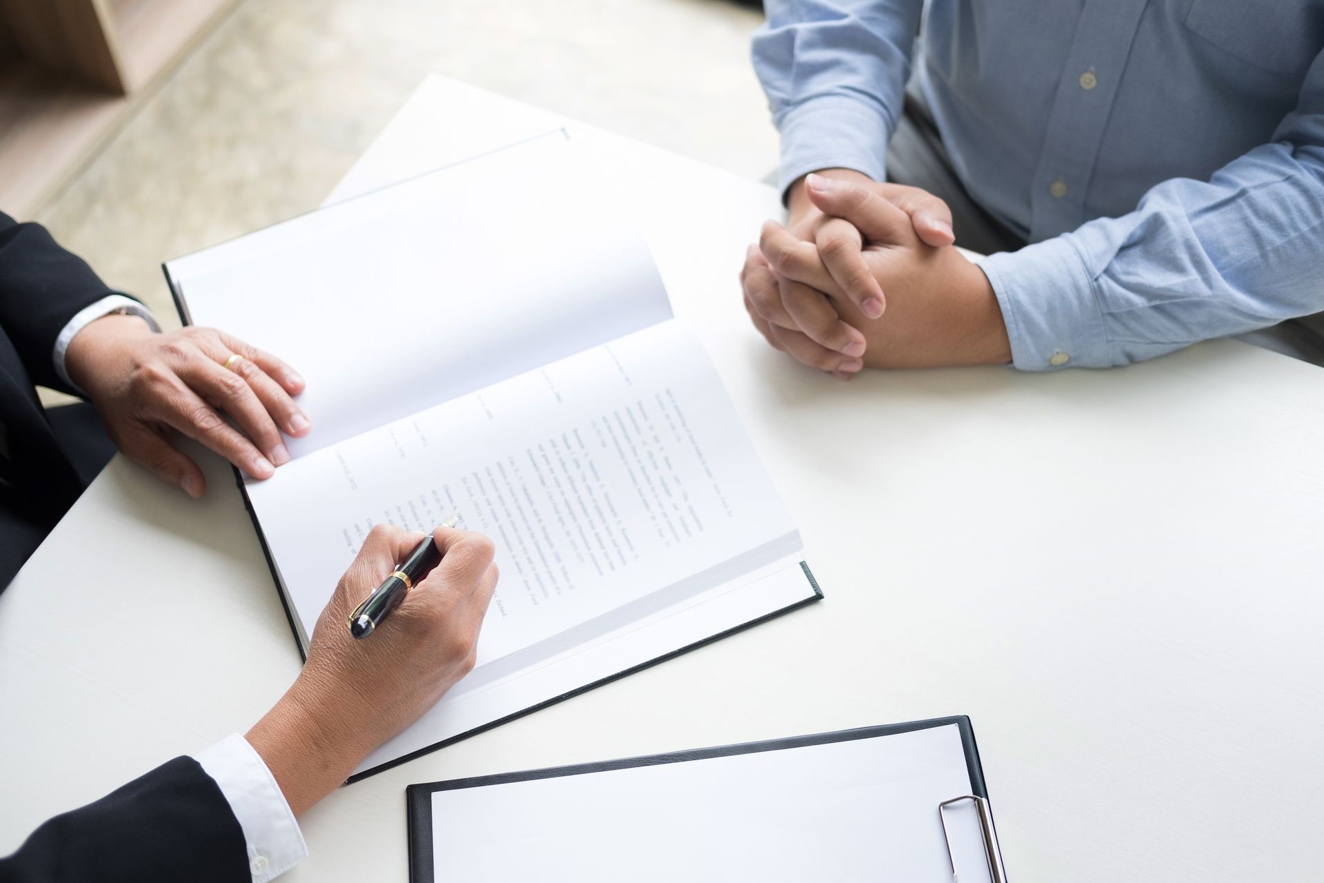 Une personne signe un document de l'autre côté d'une table, les mains jointes.