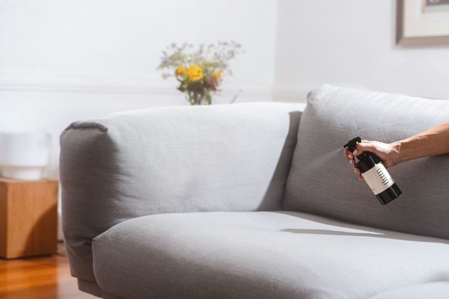 Hand holding a spray bottle beside a gray couch in a bright living room