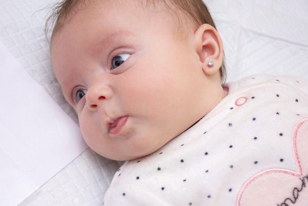 Baby with pierced ears, lying on white, looking to the side with a slightly puckered mouth.