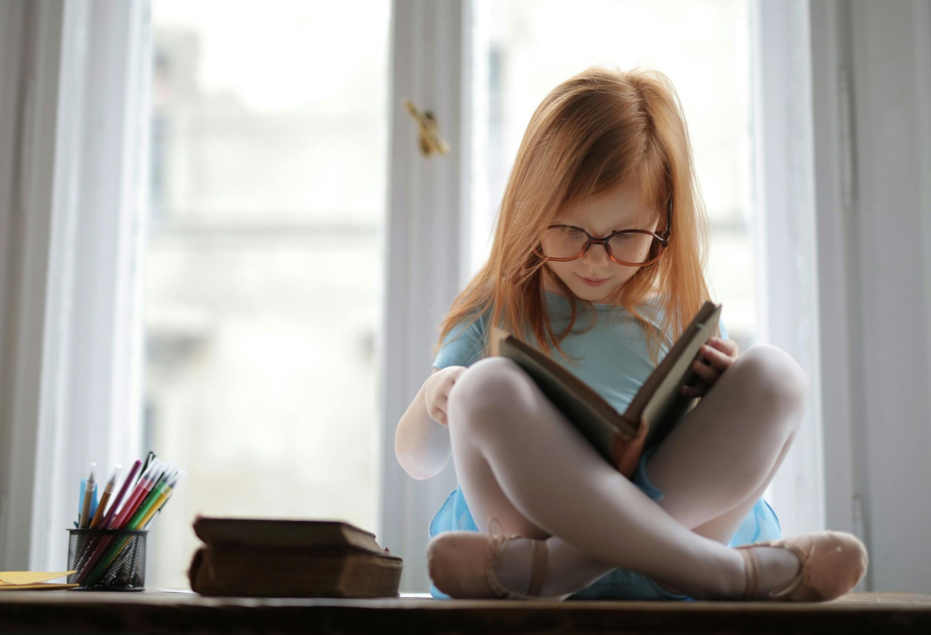 Young girl with red hair, wearing glasses, reads a book by a window.