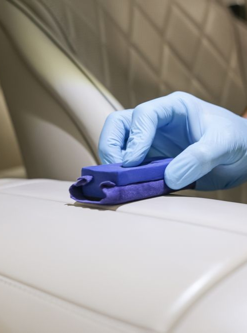 Blue-gloved hand cleaning a beige leather car seat with a purple cloth