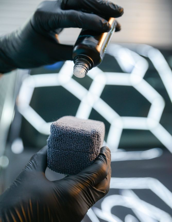 Gloved hands spray coating onto a small 3D-printed cube in front of a patterned car grille