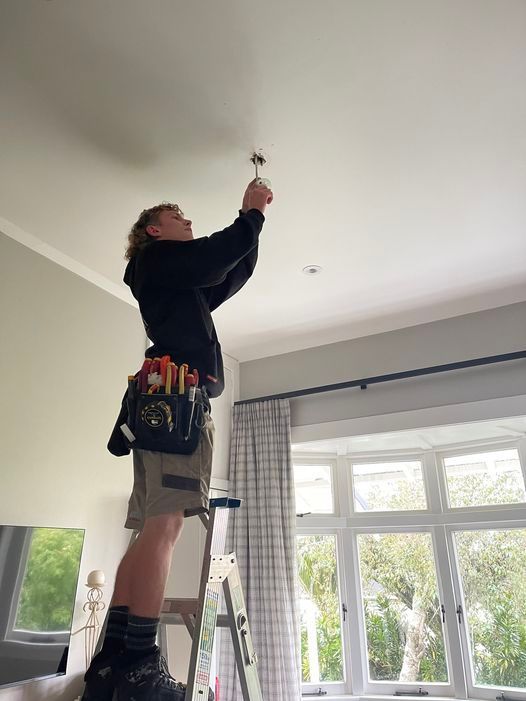 A man is standing on a ladder in a living room fixing a light fixture.