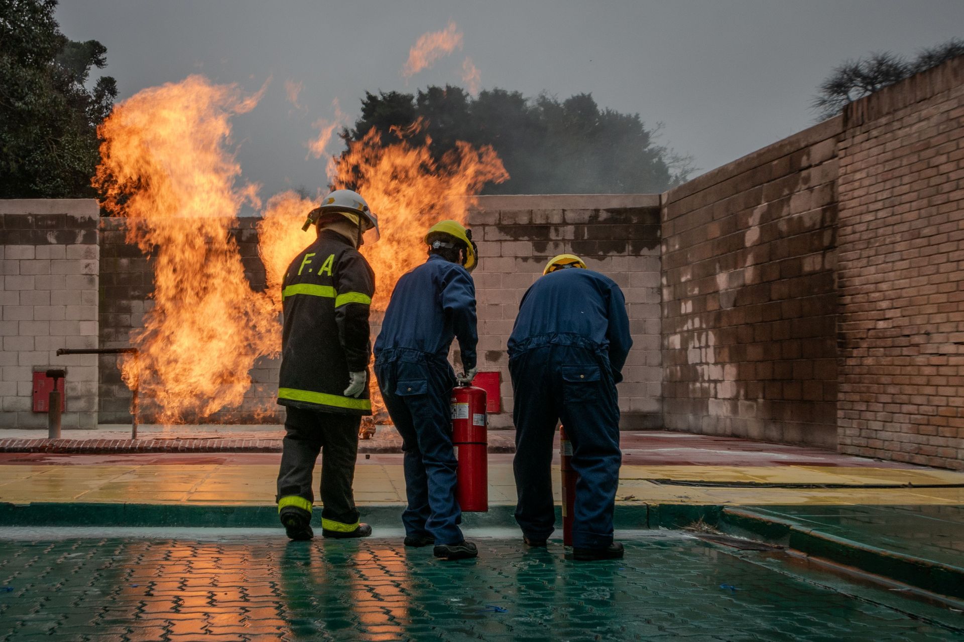Three firefighters stand before a wall of flames in a wet training yard.
