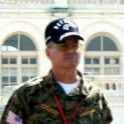 Man in camouflage shirt and cap; in front of a building with arched windows.