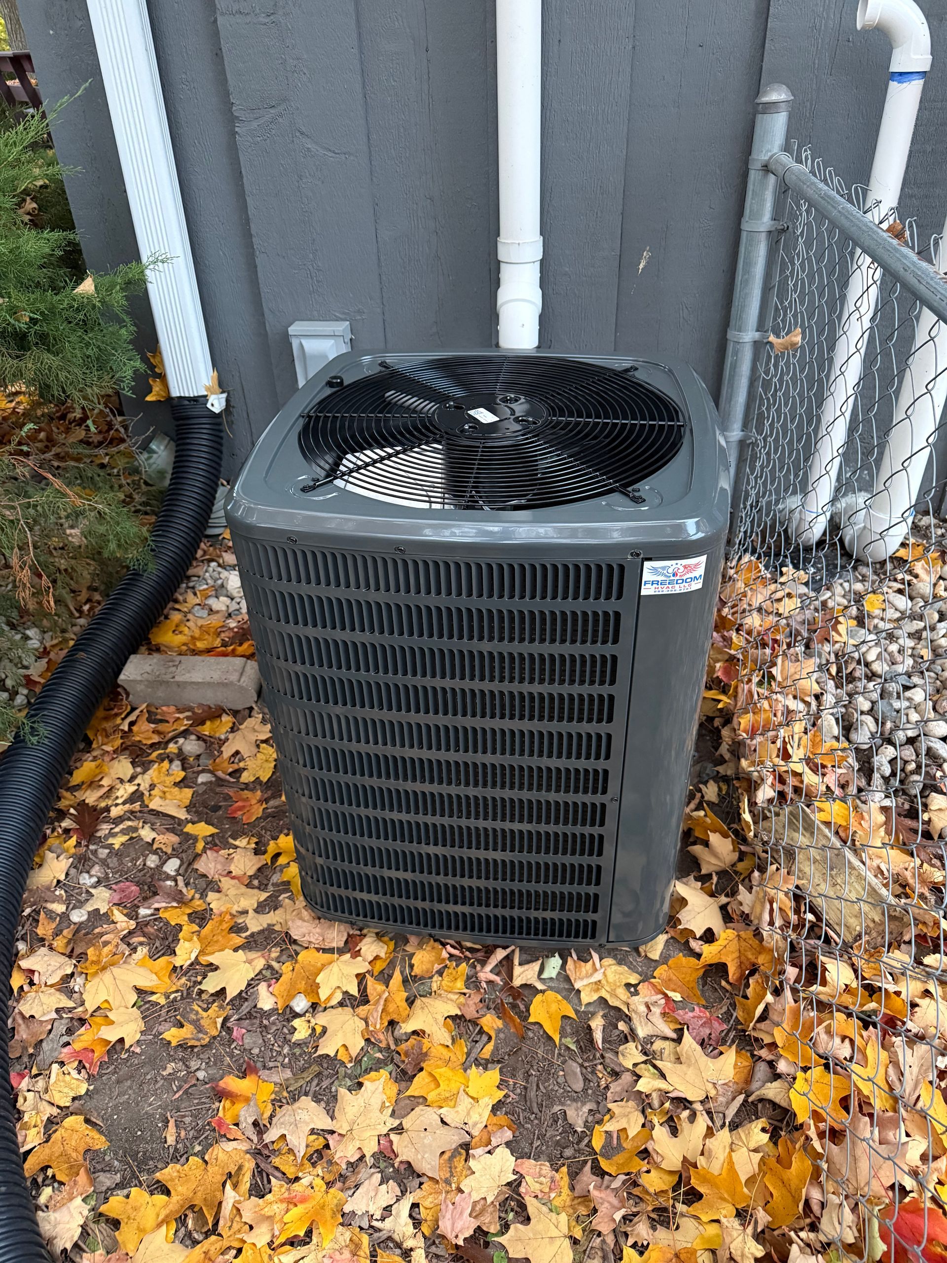 A gray HVAC outdoor condenser unit sitting on gravel surrounded by fallen autumn leaves against a building wall.