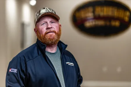 Man in a camo cap and dark jacket standing indoors with a blurred sign in the background