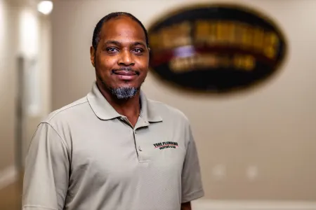 Man standing indoors in a beige polo shirt, smiling in front of a blurred wall sign.