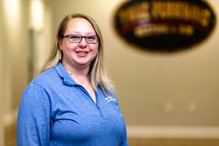Smiling person in a blue shirt standing indoors with a blurred sign in the background