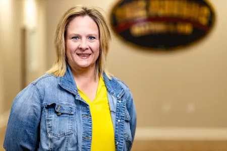Woman in a yellow shirt and denim jacket smiling indoors with a blurred sign in the background