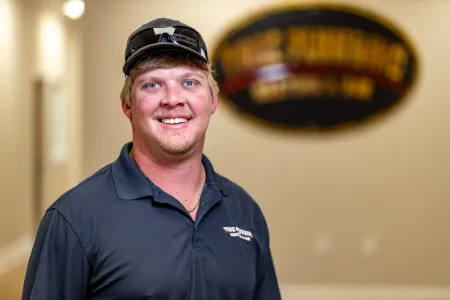 Smiling man in a dark polo and cap standing indoors with a blurred logo sign behind him