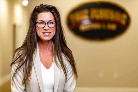 Smiling woman with glasses in a beige blazer standing indoors near a blurred sign