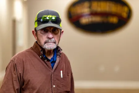Man in a brown shirt and cap stands indoors before a blurred wall sign.