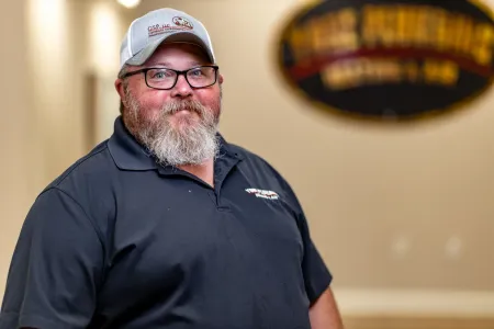 Bearded man in a cap and navy polo standing indoors with a blurred sign in the background