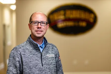Man standing indoors in a gray patterned pullover, with a blurred wall sign in the background