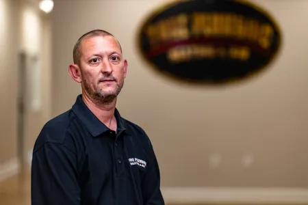 Man standing indoors in a black shirt, with a blurred office sign in the background
