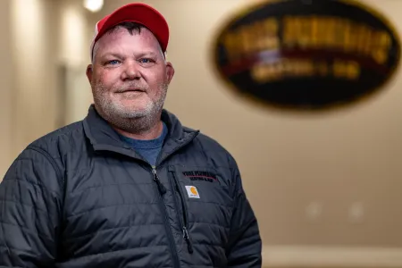 Smiling man in a red cap and black jacket standing indoors with a blurred wall sign behind him
