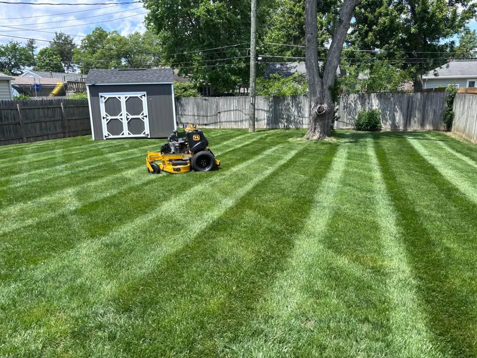 A person on a yellow riding lawnmower cuts a striped pattern into a green lawn in a backyard.