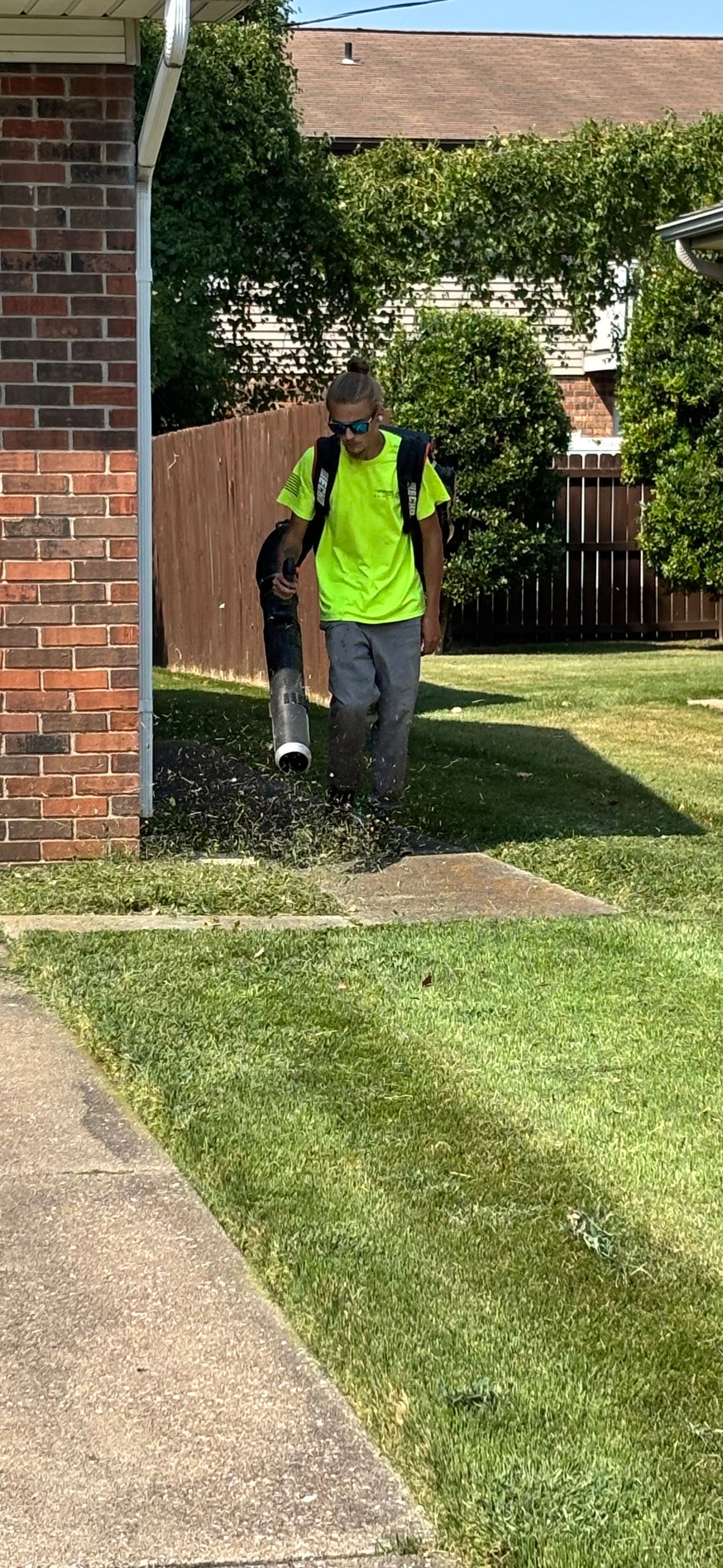 Teenager with a leaf blower on a concrete driveway, wearing sunglasses, yellow shirt, and gray pants.