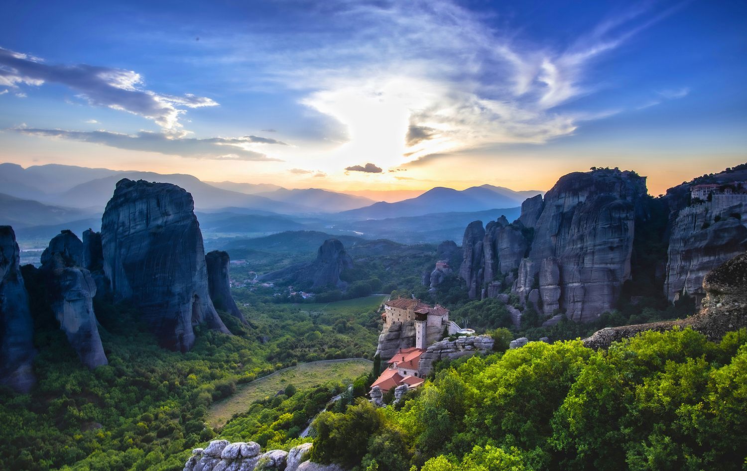 A monastery sits on a high rock formation overlooking a lush valley at sunrise under a blue and golden sky in Meteora.