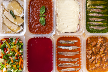 Piatti pronti assortiti in contenitori di plastica, tra cui verdure, salse, pasta e pietanze impanate.