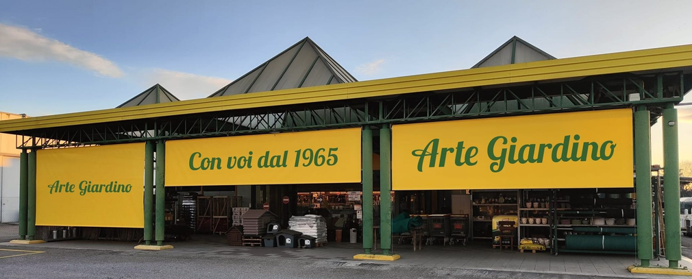 L'esterno di un centro giardinaggio Arte Giardino con striscioni gialli, pilastri di sostegno verdi e un tetto di tegole grigie.