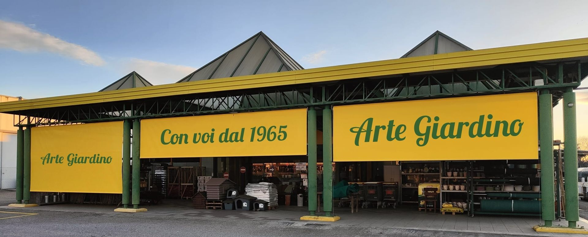 L'esterno di un centro giardinaggio Arte Giardino con striscioni gialli, pilastri di sostegno verdi e un tetto di tegole grigie.