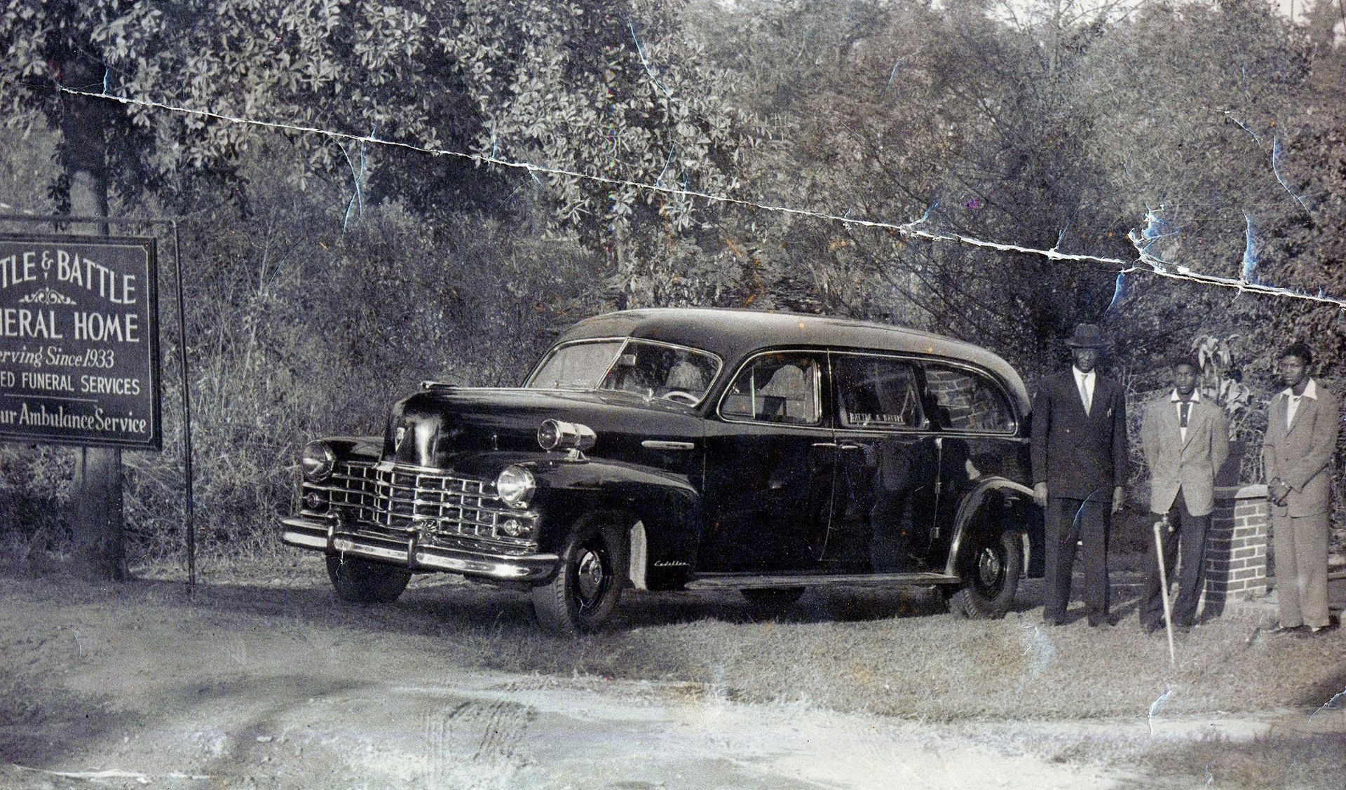 A black vintage hearse parked in front of a funeral home sign, with three people standing to its right.