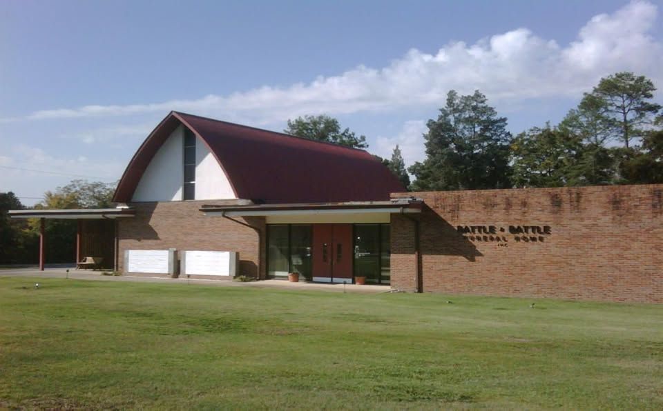A brick building with a curved red roof on a grassy lawn with trees in the background under a blue, cloudy sky.