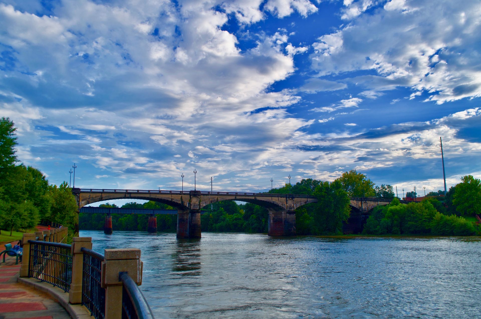 A stone arch bridge spans a wide river under a dramatic blue sky filled with textured white clouds.