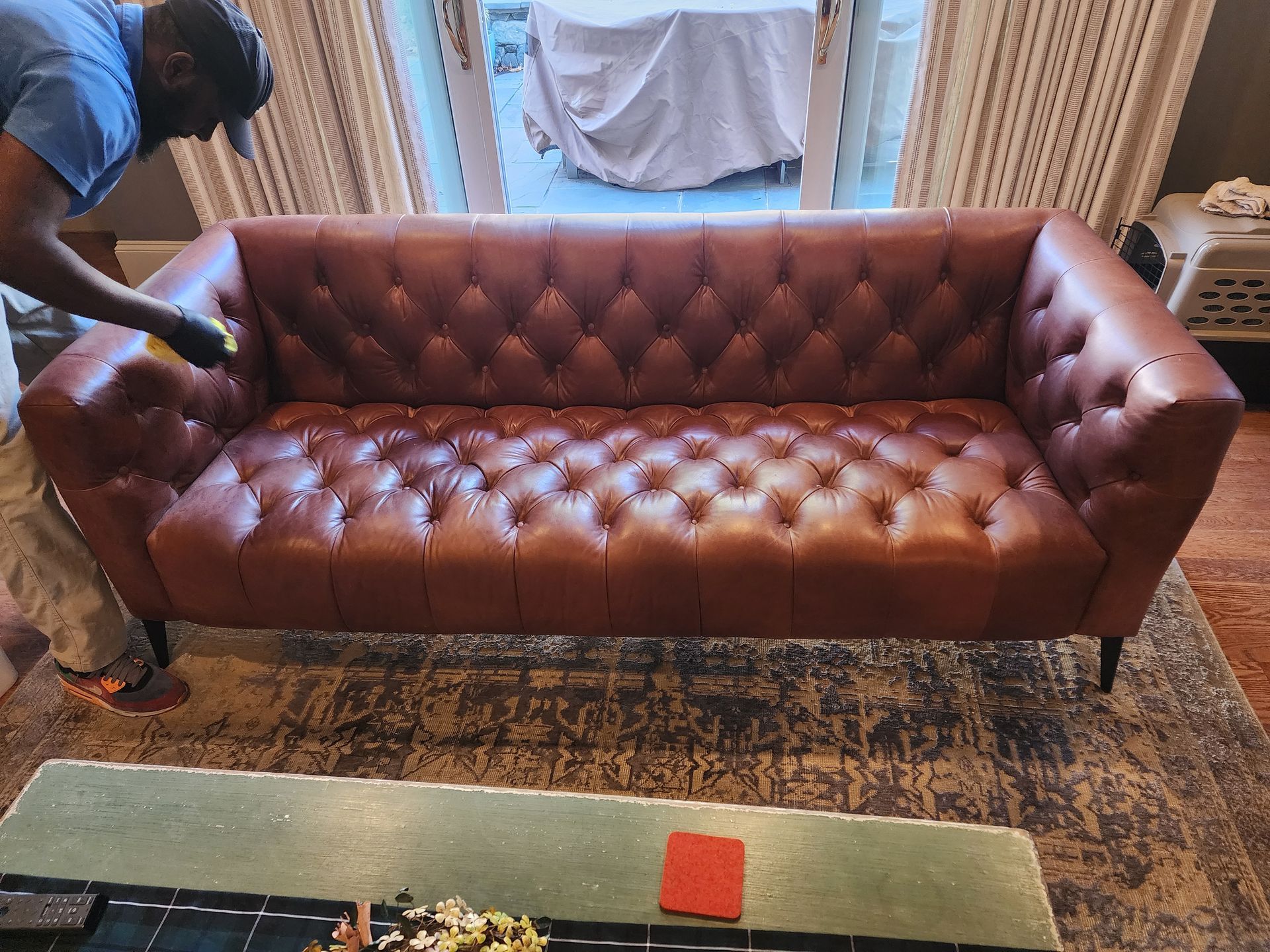 Person cleaning a brown tufted leather sofa in a living room with a patterned rug.