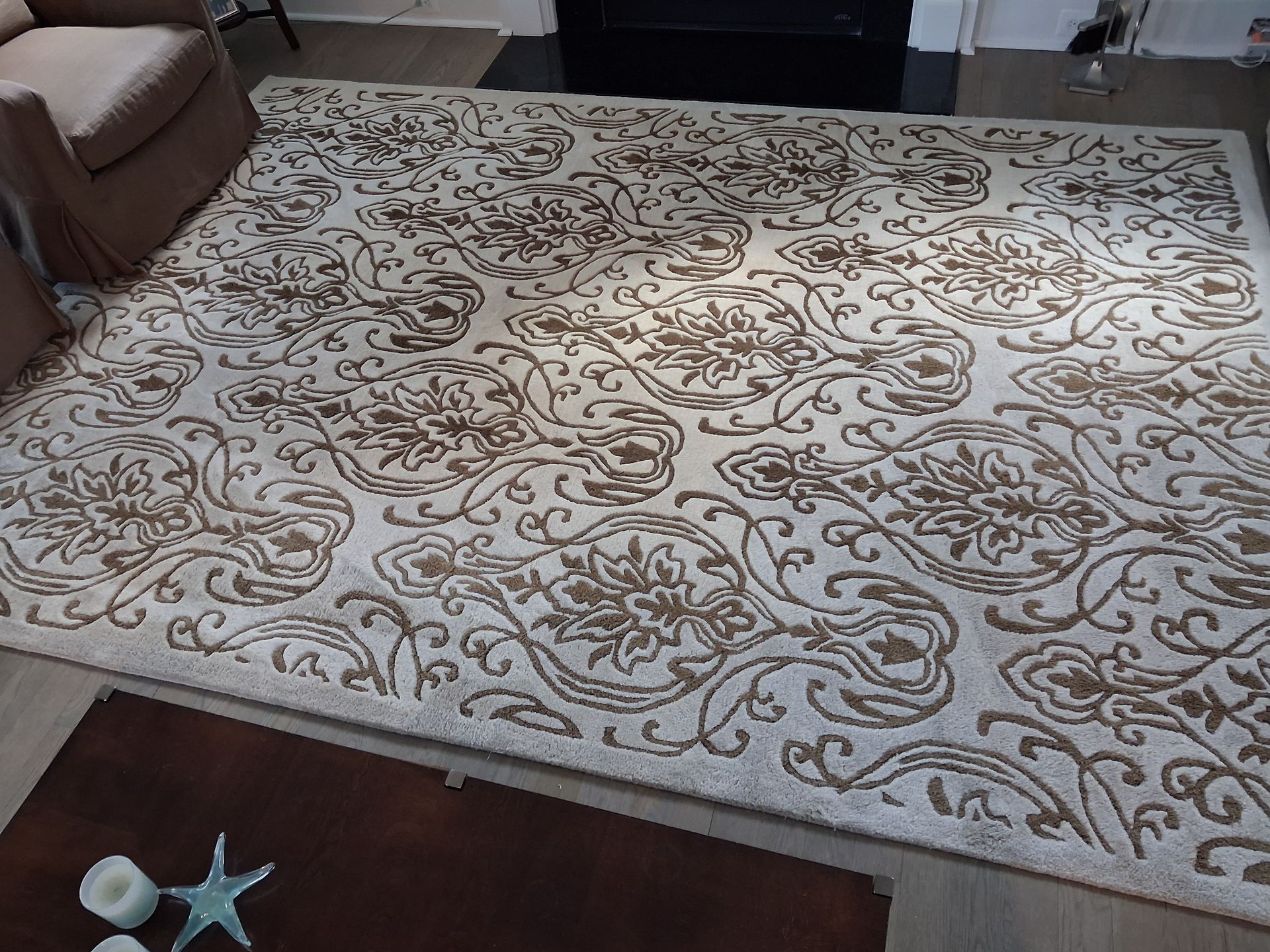 Large cream rug with brown floral swirls on a dark floor, with a small starfish and shell decoration in the corner