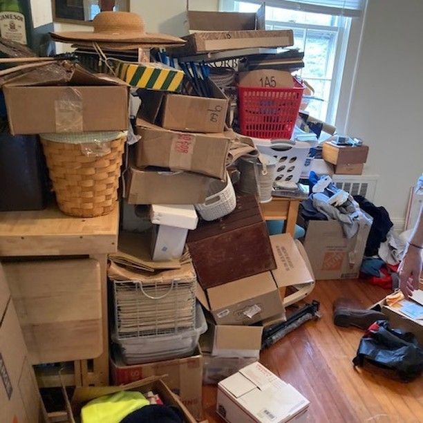 Cluttered room with stacked cardboard boxes, baskets, and miscellaneous items near a window.