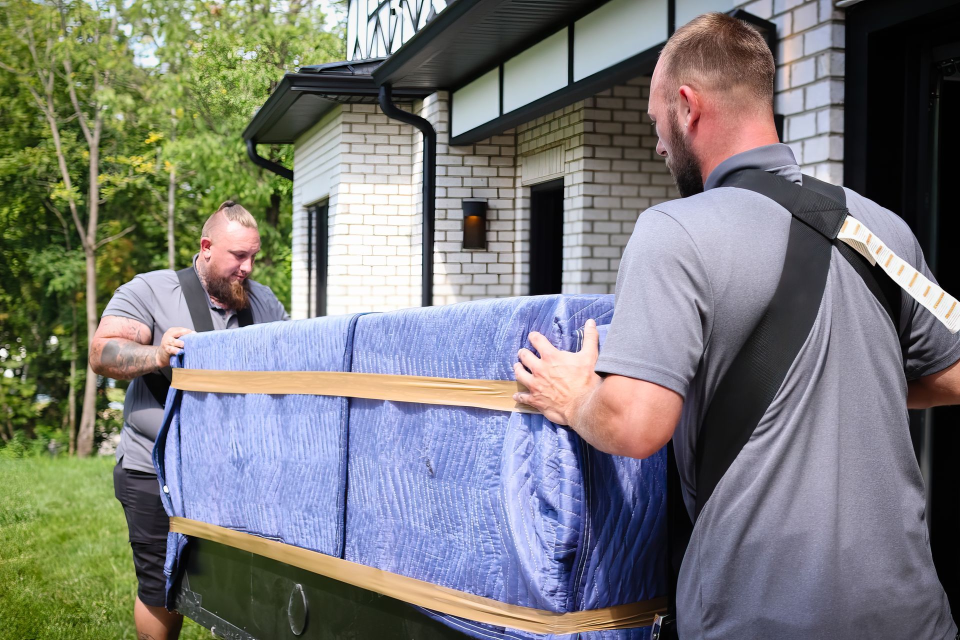 Two movers carry a large, wrapped object out of a brick building onto a grassy area.