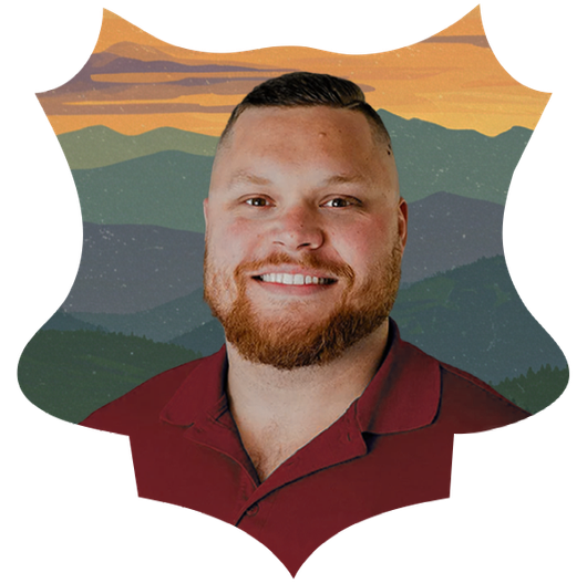 Man with a red beard smiles, set against a mountain landscape illustration, wearing a burgundy shirt.