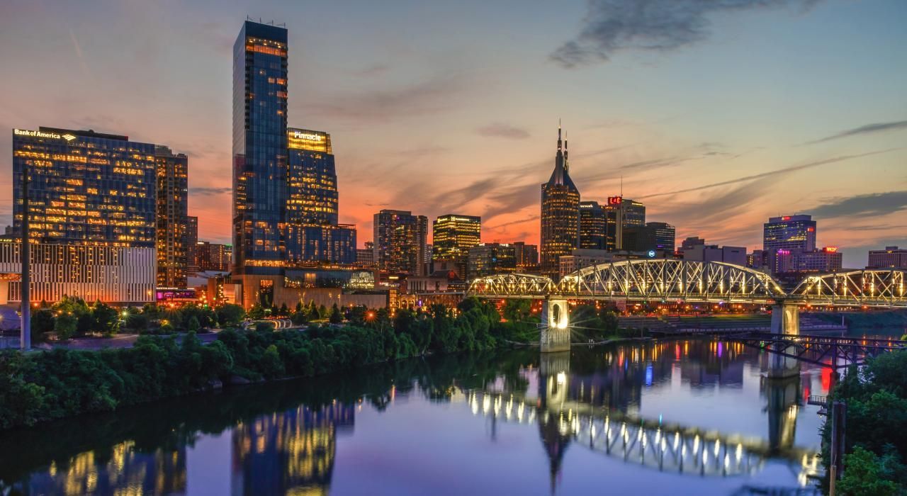Nashville skyline at dusk with river reflection. Buildings and bridge lit up against a colorful sky.