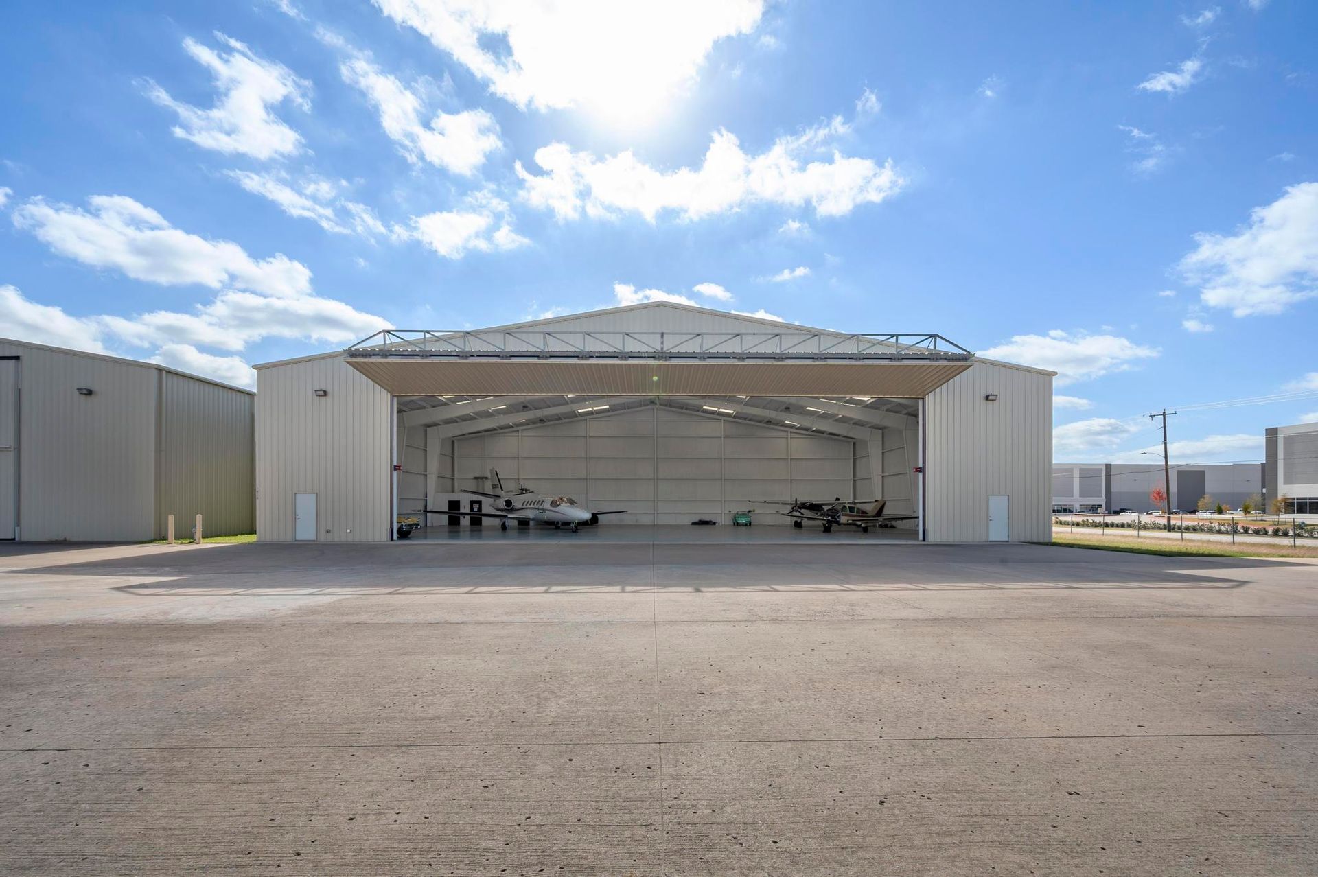 Open-sided hangar with parked aircraft under a bright blue sky, viewed across a gravel lot