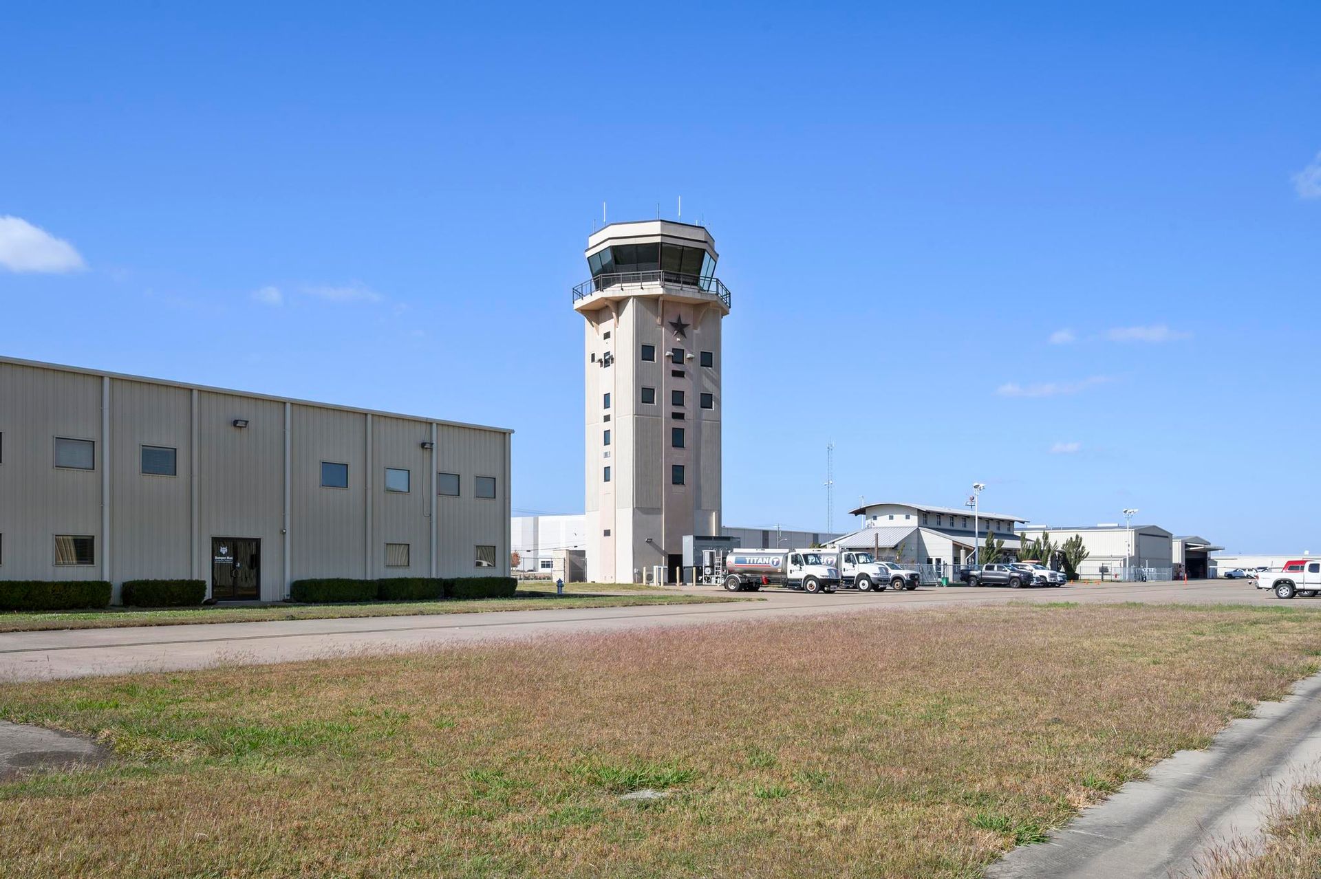 Airport control tower beside hangars on a sunny day, with a grassy foreground and blue sky.