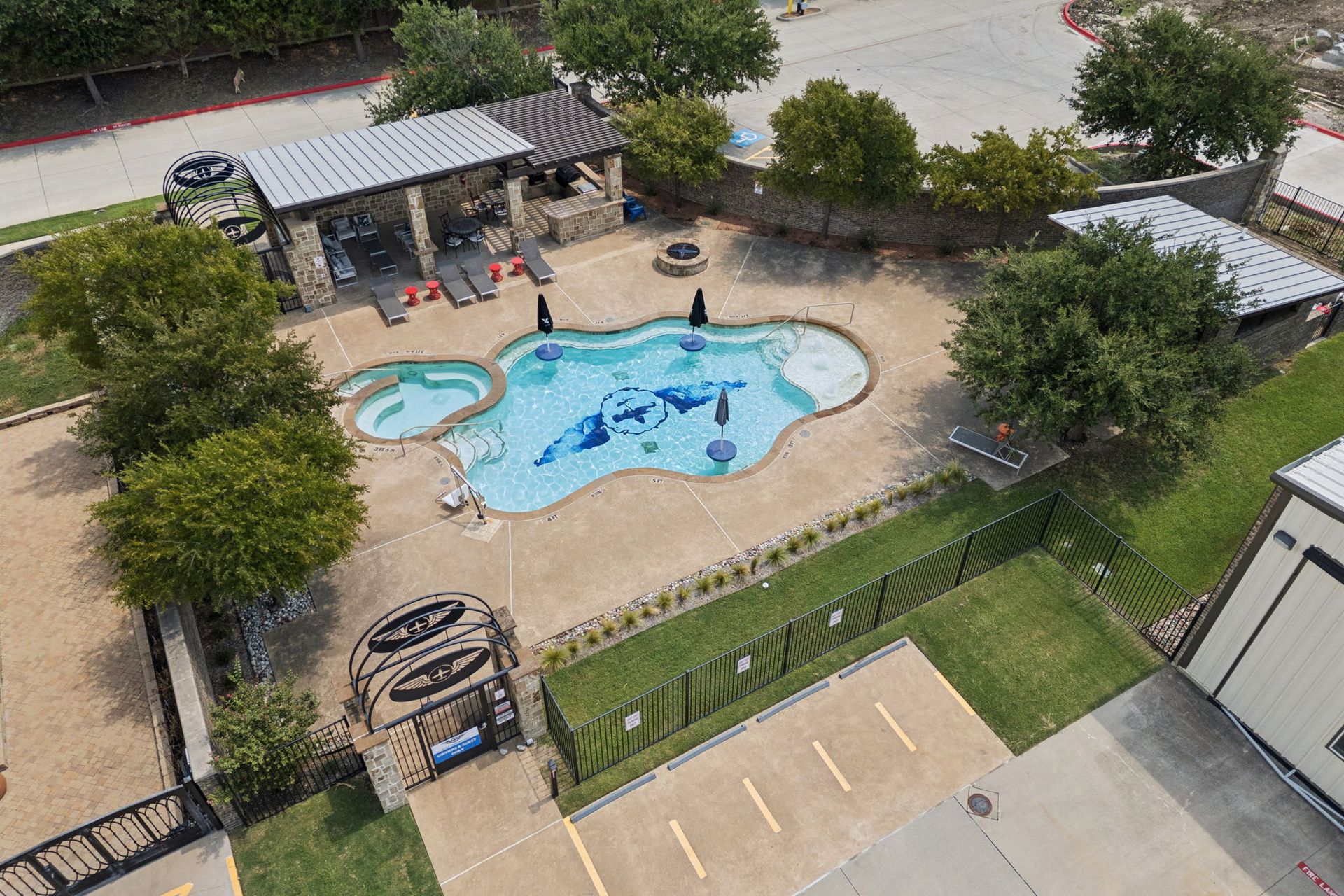 Aerial view of a small outdoor pool area with a splash pad, shaded seating, and surrounding fence.