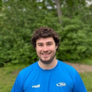 Man with curly brown hair, wearing a blue shirt, smiling in front of a green leafy background.